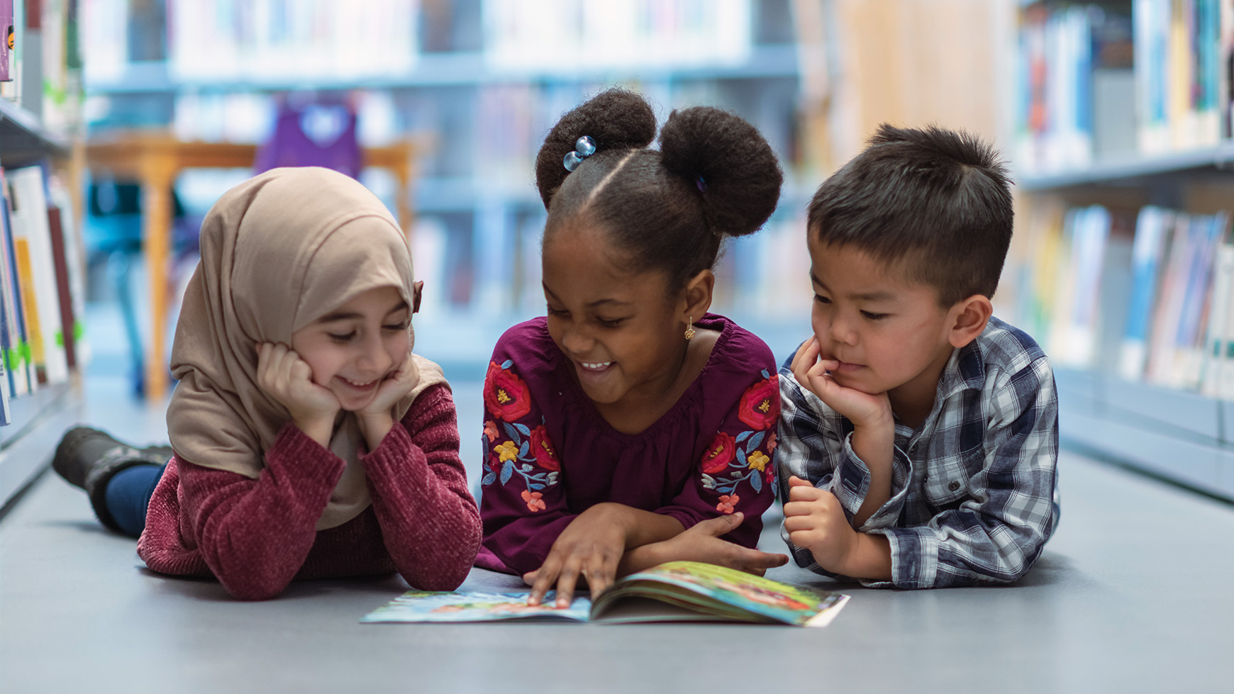 Three (3) kids who are friends are laying on the floor in between bookshelves in the library. They are reading a book together. They are all smiling and enjoying their afternoon.
